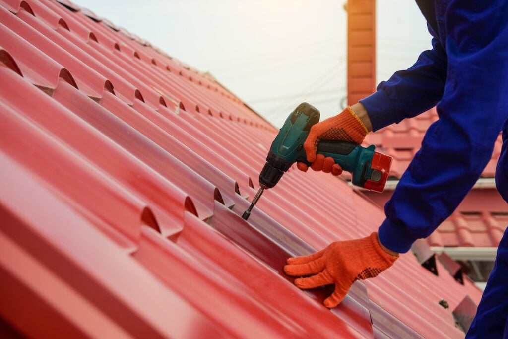 Close up hands in protective gloves of young man worker who fix a metal tile roof with drill.
