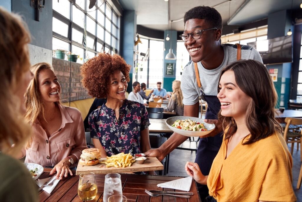 Waiter Serving Group Of Friends Meeting For Drinks And Food In Restaurant